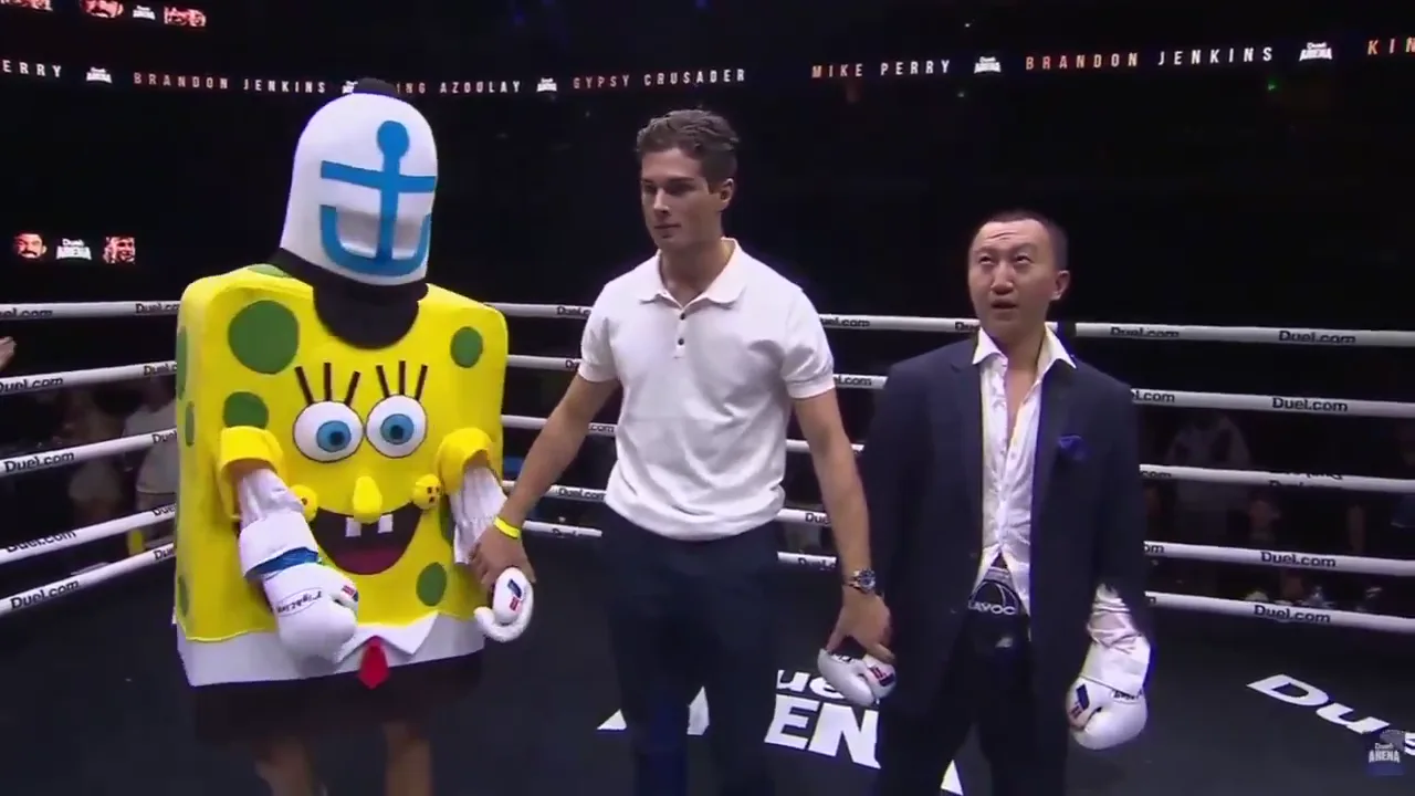 A performer in a yellow sponge costume with an anchor hat and red tie stands in the Duel Arena ring beside a man in a white polo shirt and a man in a navy suit wearing boxing gloves